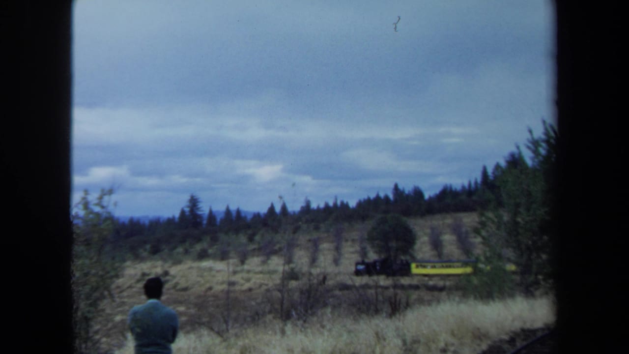 A man in a green sweater watches a small yellow train move across a dry California field in 1970 home-movie footage.