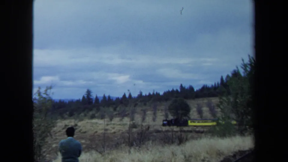 A man in a green sweater watches a small yellow train move across a dry California field in 1970 home-movie footage.