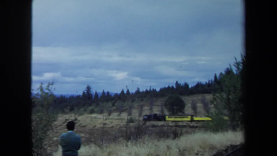 The train continues across the open field as the man watches from the foreground, framed by 8mm gate edges and cloudy sky.
