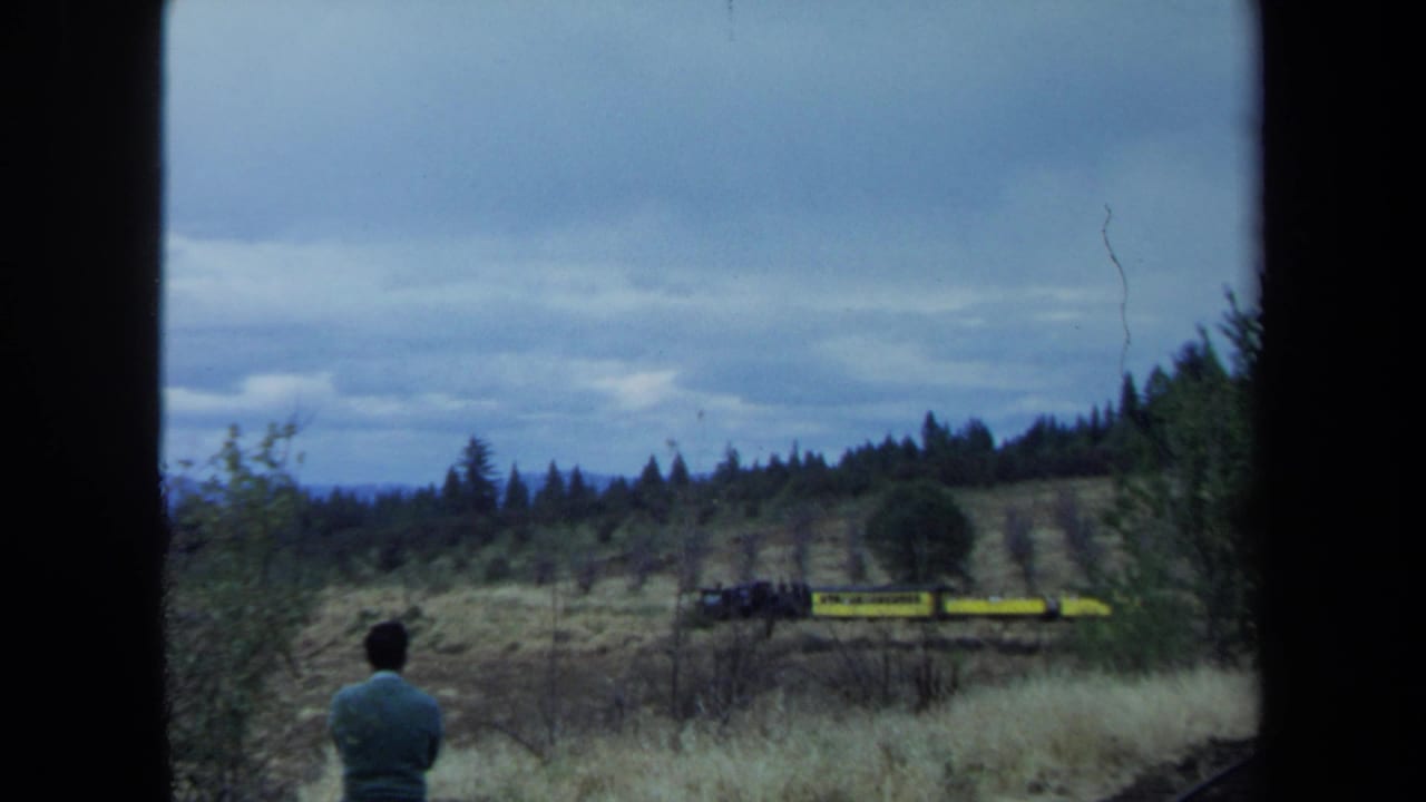 A man seen from behind watches a yellow freight train pass through a wooded California hillside in 1970 home-movie footage.