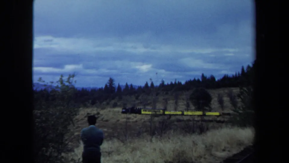 Home-movie view of a man facing a distant yellow freight train, with dry grass, trees, and cloudy hills in California.