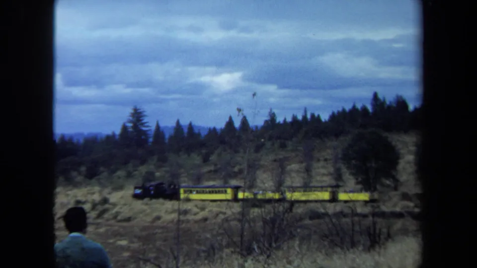 A distant figure in a blue sweater faces a passing yellow train in a rural California landscape on 8mm film.