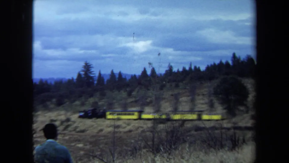 Soft 8mm frame of a lone observer and a yellow train crossing a brushy California hillside beneath heavy clouds.