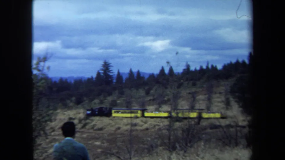 1970 California 8mm frame of a man seen from behind as a small yellow train moves across a wooded hillside under cloudy sky.