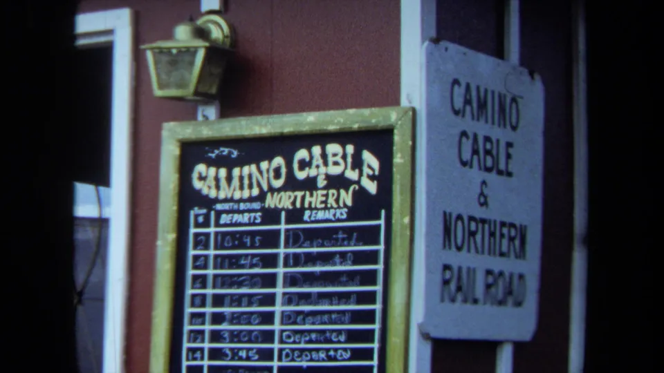 1970 California 8mm frame showing a Camino Cable & Northern Railroad chalkboard schedule and adjacent wall sign near a red station wall.