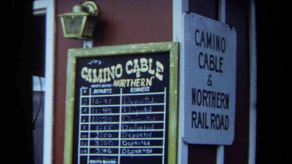 1970 California 8mm frame showing Camino Cable and Northern Railroad signage, a posted departure board, red siding, and a wall lantern.
