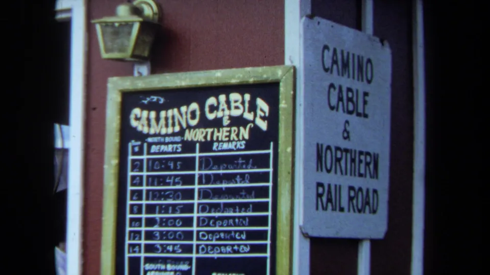 1970 California 8mm frame showing the station schedule board, handwritten departure times, lantern, and Camino Cable and Northern Railroad wall sign.