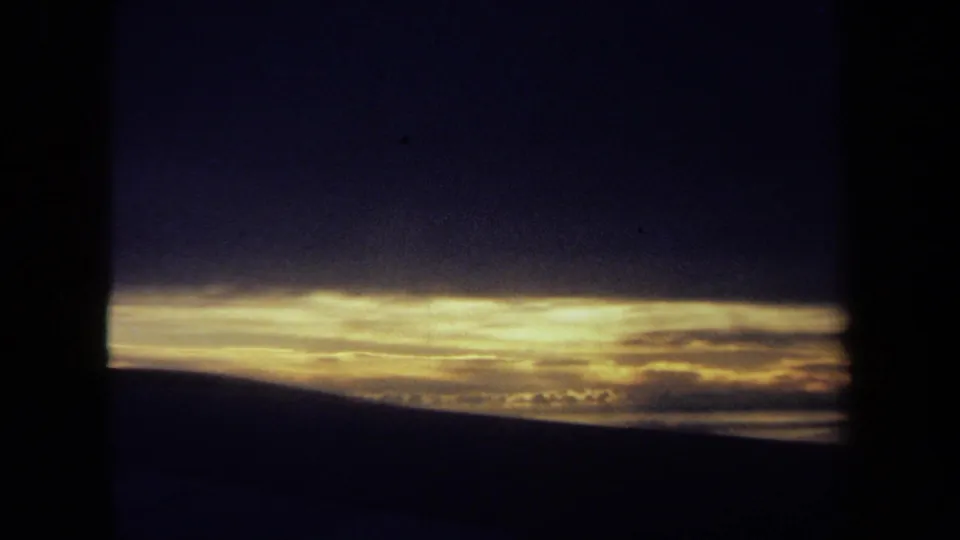 Grainy 1970 California 8mm sunset view with dark clouds, a bright golden band of light, and a low landscape horizon.