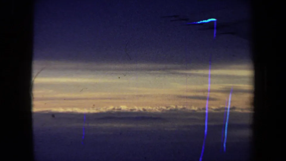 The frame shows a low-light airplane-window view of sunset clouds, with a dark window divider and blue-purple film streaks visible in the 8mm scan.