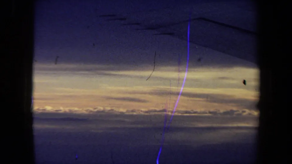 A faint airplane wing silhouette appears against a blue sunset sky and warm cloud horizon, framed by the dark window edge in the 1970 California travel footage.