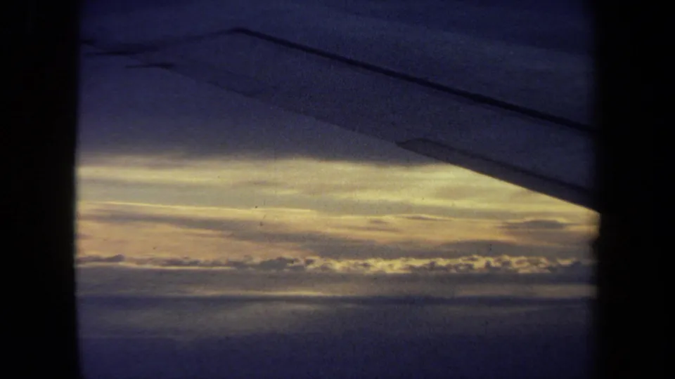 1970 California home-movie frame showing an aircraft wing edge above a glowing cloud horizon through a passenger window.