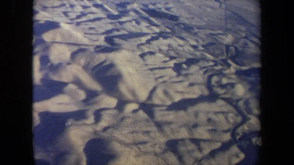 1970 California aerial view from a passenger airplane window, showing pale rugged terrain with shadowed ridges below.