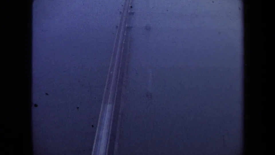 Blue-tinted 8mm frame showing the bridge or causeway descending through the aerial view over water, with dust and gate edges visible.