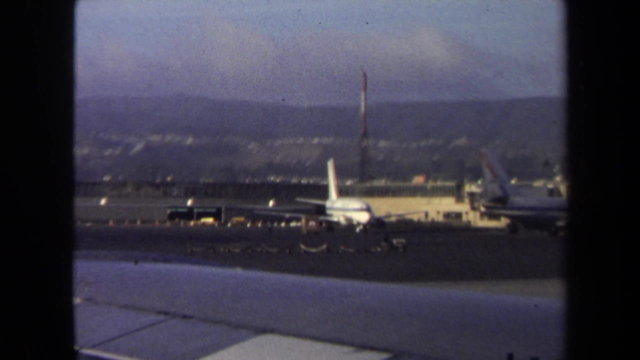 1970 Washington, DC airport tarmac view with parked airliners, a luggage tram crossing the pavement, and hazy hills beyond.