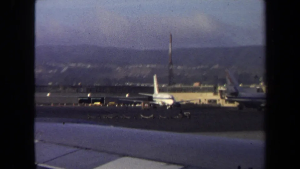 1970 Washington, DC airport tarmac view with parked airliners, a luggage tram crossing the pavement, and hazy hills beyond.