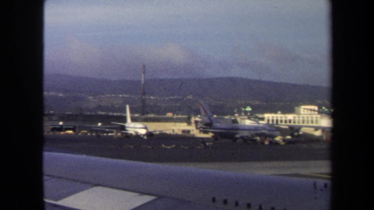 Airliners parked near airport buildings with runway pavement and hills in the background.