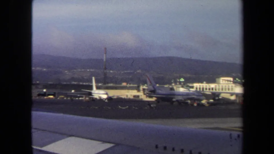 Passenger-window view of a Washington, DC airport scene in 1970, showing airliners, terminal structures, runway pavement, and distant hills.