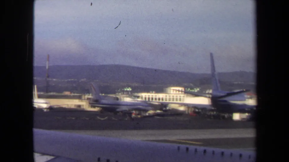 1970 Washington, DC tarmac view with a parked commercial aircraft beside terminal buildings and distant hills under a hazy sky.