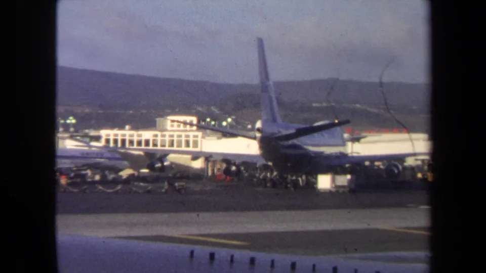 Close tarmac view from the 1970 Washington, DC clip showing an airliner tail, terminal buildings, service area, runway, and hazy hills.