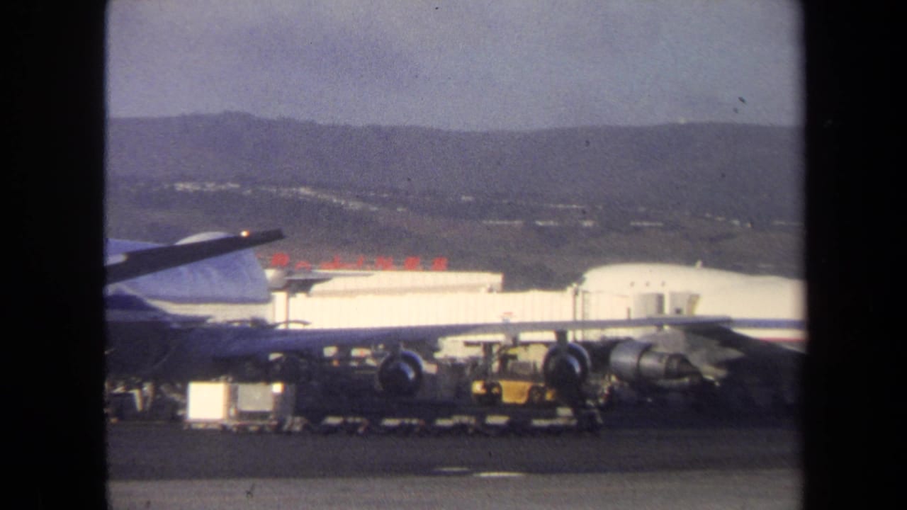 1970 8mm frame showing a jet on an airport ramp with ground equipment, low buildings, and hills in the distance.