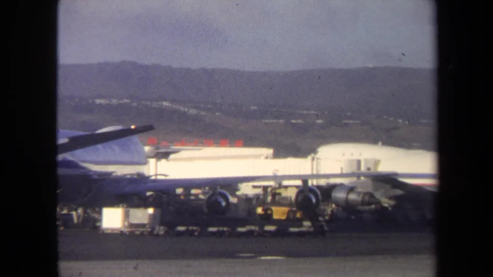 1970 8mm frame showing a jet on an airport ramp with ground equipment, low buildings, and hills in the distance.