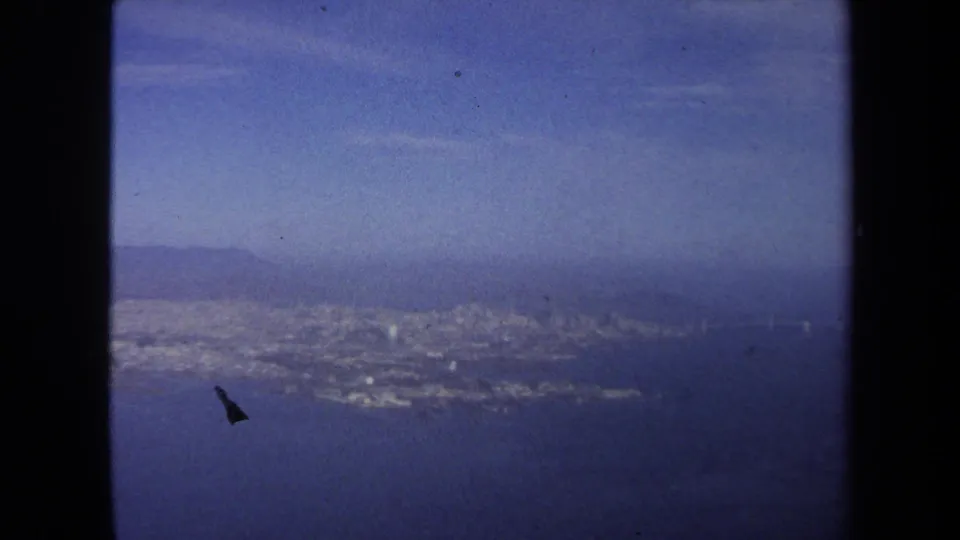 1970 8mm airplane-window view looking over blue water toward a hazy coastal city and low hills.