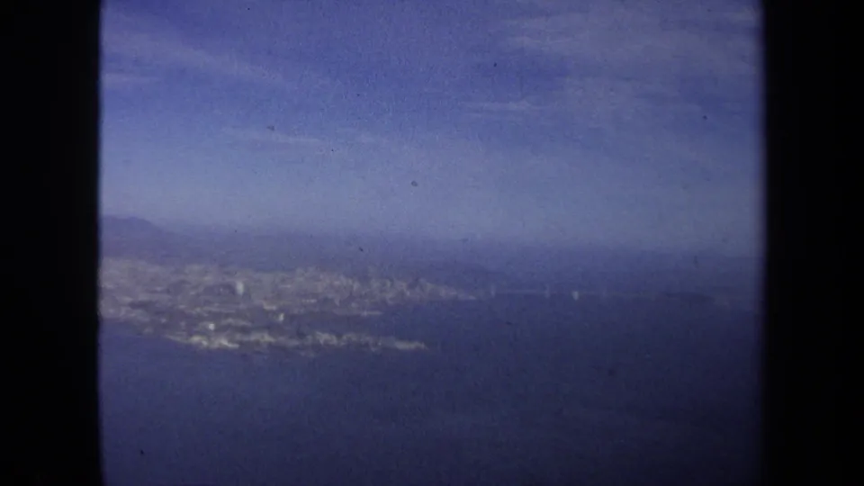 1970 8mm frame with a soft airplane-window view of coastline, water, shoreline development, and distant hills.