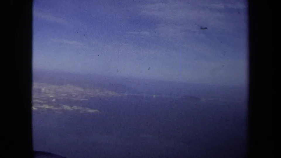 1970 8mm airplane-window frame showing blue water, a faint coastline, distant land, and soft atmospheric haze.