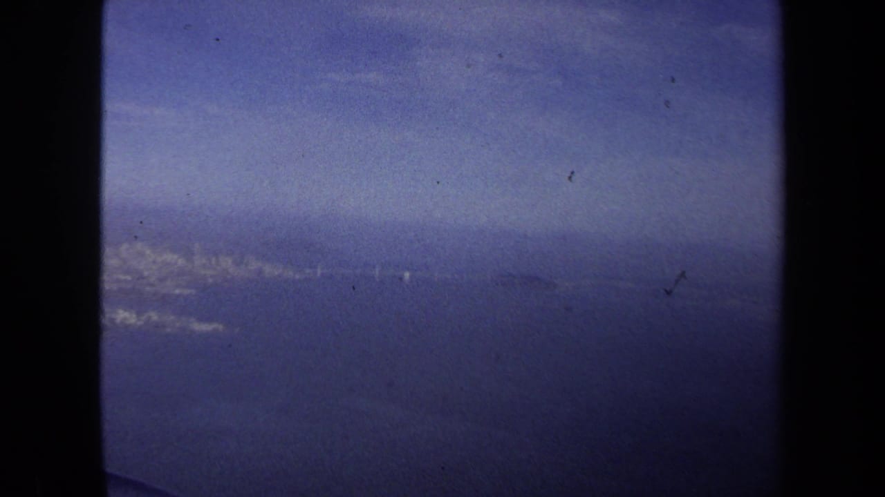 1970 8mm home-movie frame from a Washington DC travel reel showing blue sky and distant terrain through an airplane window.