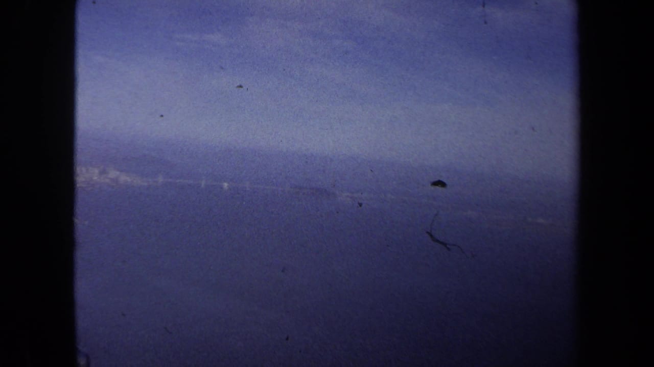 Passenger-window view from the 1970 travel footage, with a distant coastline or waterline softened by haze and film grain.