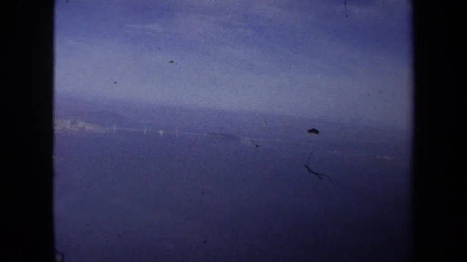 Passenger-window view from the 1970 travel footage, with a distant coastline or waterline softened by haze and film grain.