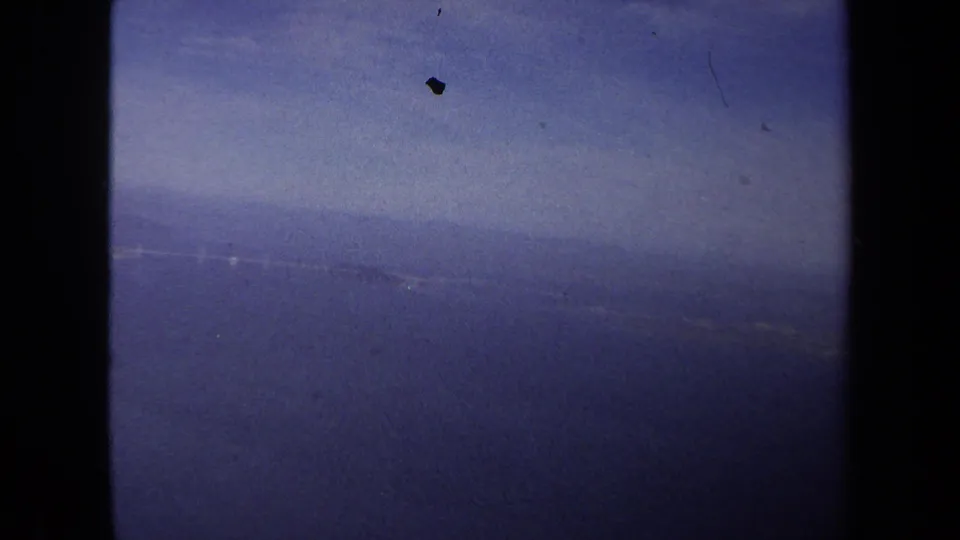 1970 8mm aerial travel frame showing water, faint coastline, and sky through the aircraft-window opening.