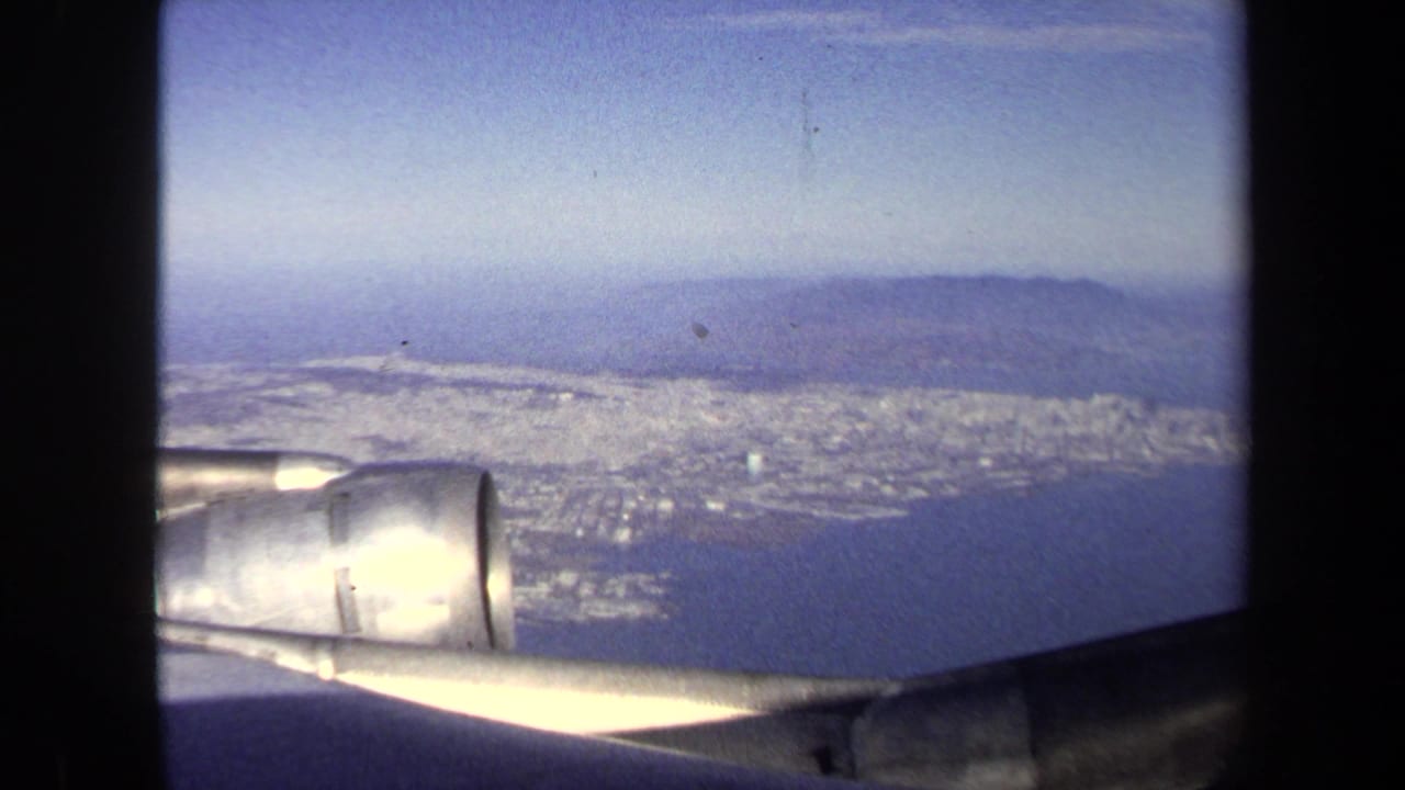 1970 8mm passenger-window view showing an airplane wing and engine above a coastal city bordered by water and hazy hills.