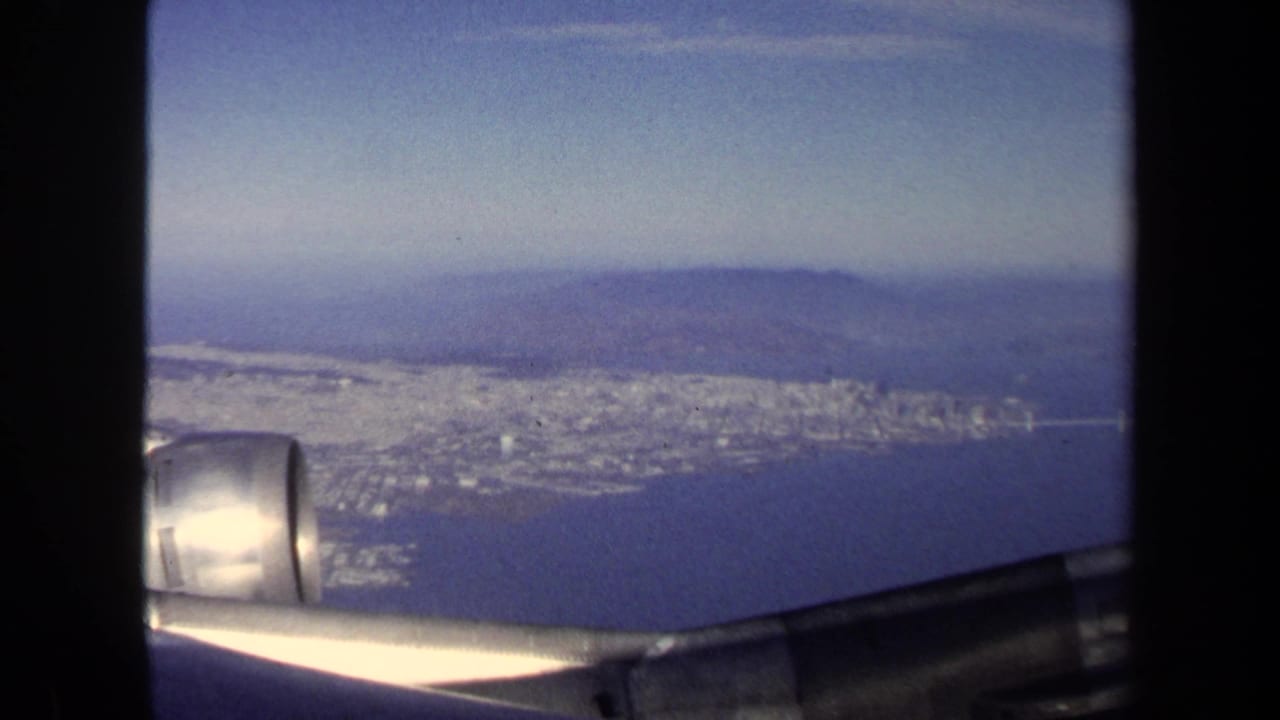 Aerial view through an airplane window with wing, engine, city shoreline, and hills.