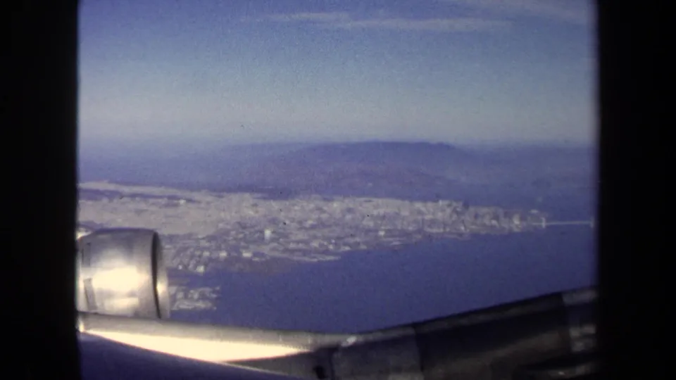 A passenger-window aerial from the 1970 8mm clip shows the aircraft wing over a bright coastal urban shoreline and distant hills.