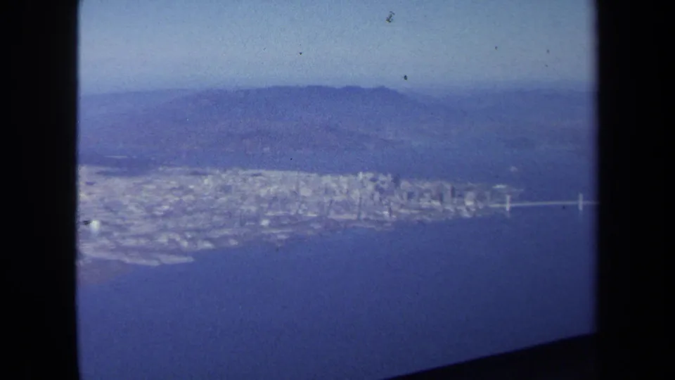 Late-frame aerial view from the 1970 8mm clip shows a dense coastal city beside blue water with hills in the distance.