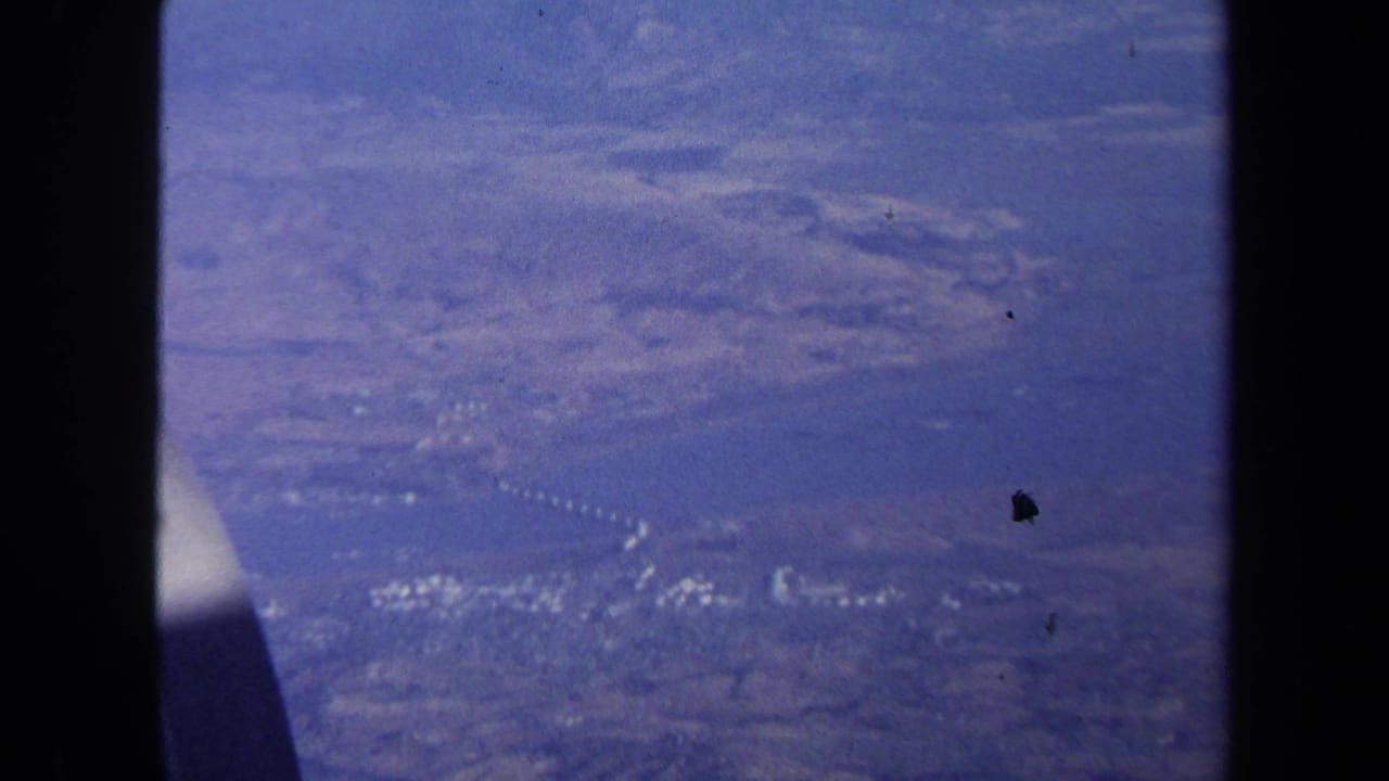 1970 aerial home-movie frame showing hazy land, water, and a thin bridge or roadway line below the aircraft.