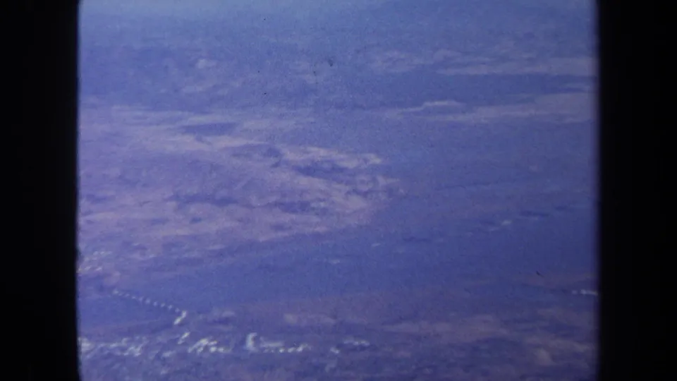 1970 8mm aerial frame with aircraft window bars, open water, shoreline shapes, pale terrain, and heavy blue film cast.