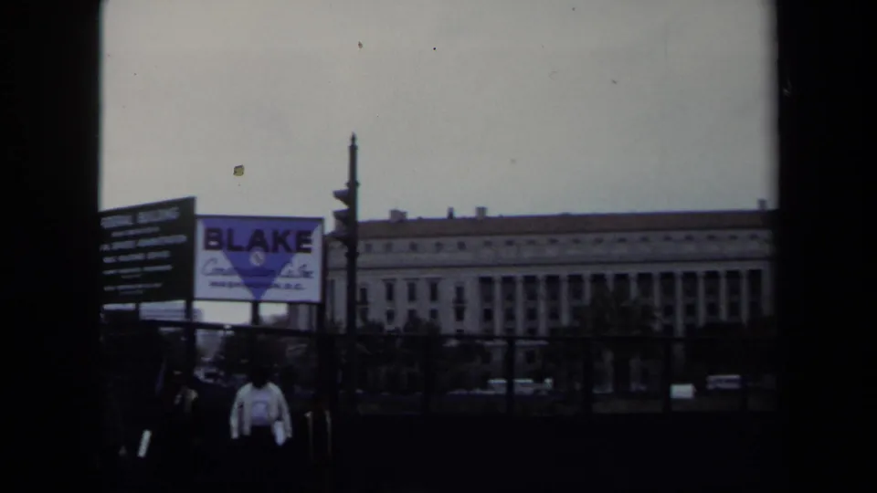 1970 Washington DC 8mm frame showing a street scene with pedestrians, signage, a billboard, and a large civic-looking building.
