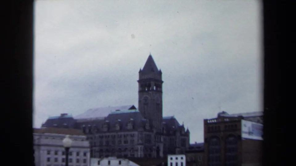 1970 8mm Washington, DC view centered on a pointed clock tower rising above lower downtown buildings under gray sky.