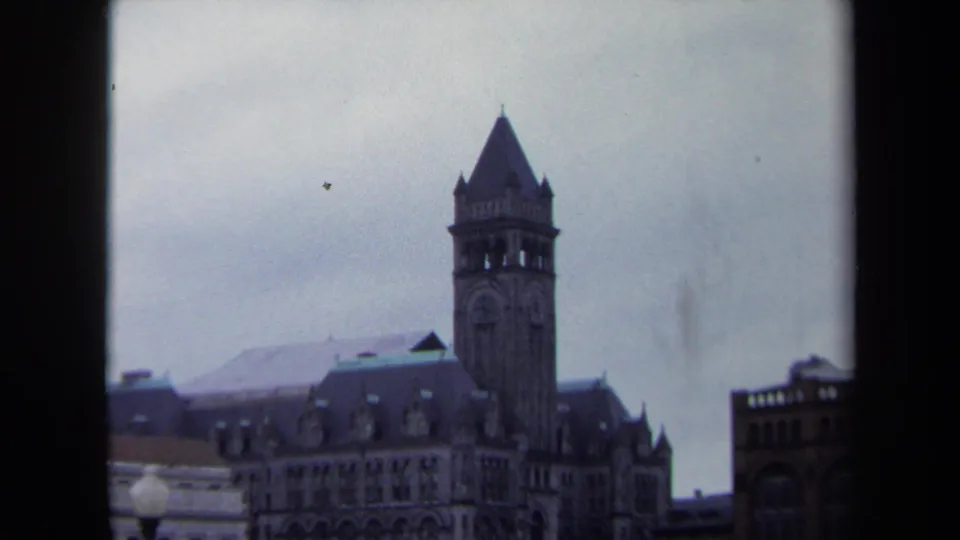 1970 8mm Washington, DC frame showing a pointed clock tower, dark-roofed building, pale facade edge, and brick building at right.