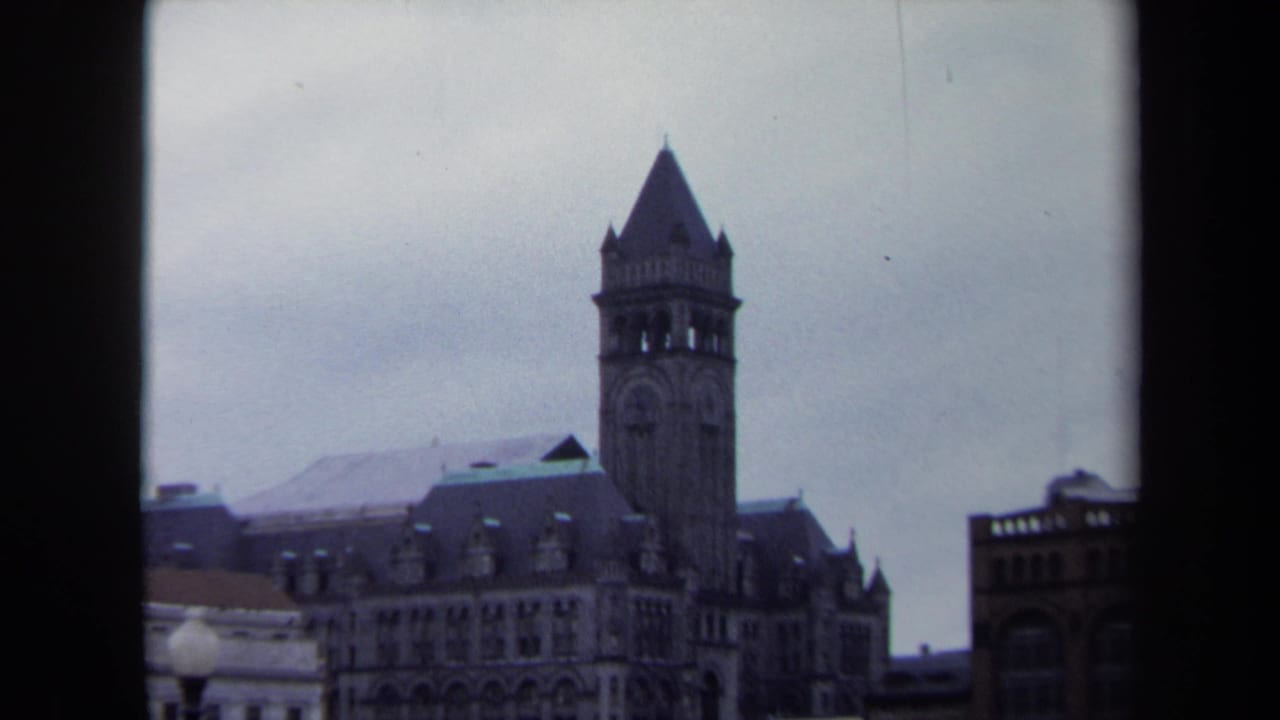 1970 Washington, DC home-movie frame showing a towered stone building beyond lower rooftops under a gray sky.