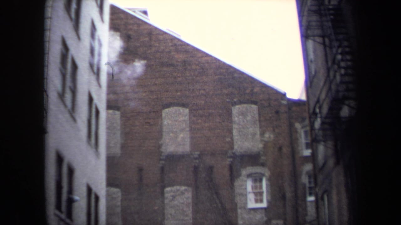 Narrow alley framed by tall buildings and a brick wall with boarded windows.