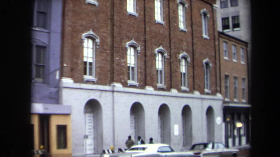 1970 Washington, DC street view showing parked cars and small pedestrian figures outside a brick building with arched openings.