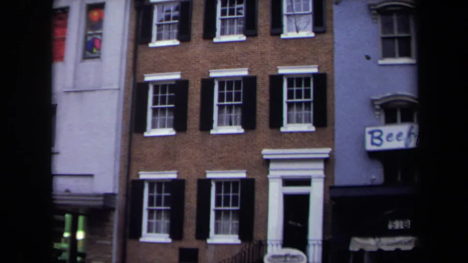 1970 Washington, DC home-movie frame showing a brick townhouse facade with black shutters, white trim, and adjacent buildings.