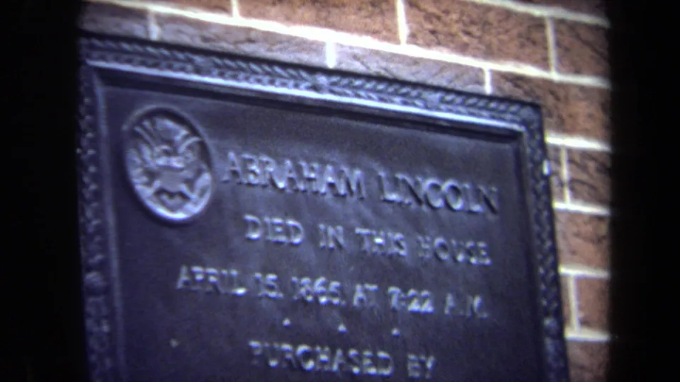 Final 8mm frame showing the Abraham Lincoln death plaque in soft focus, with raised letters and red-brown brick behind it.