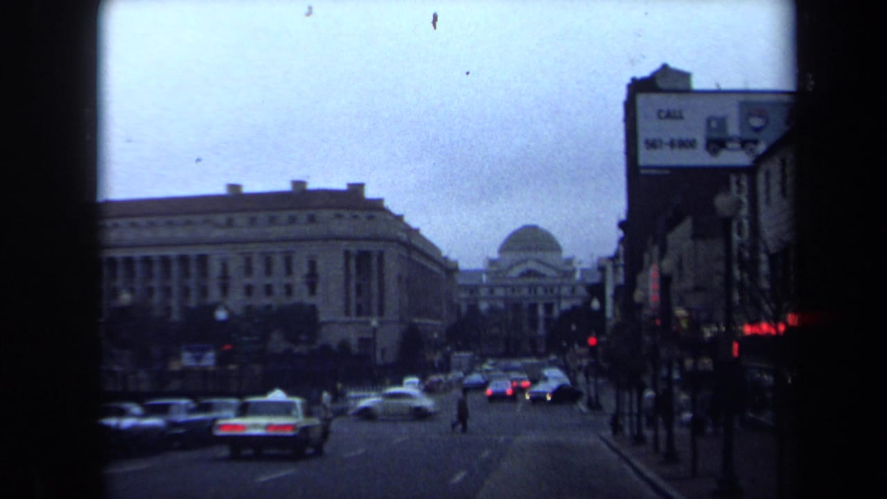 1970 Washington, DC street view with classic cars, pedestrians, traffic lights, storefronts, and a distant domed civic building.
