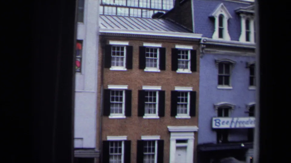 1970 Washington, DC streetscape with adjoining rowhouse facades, black shutters, white doors, dormers, and a partial storefront sign.