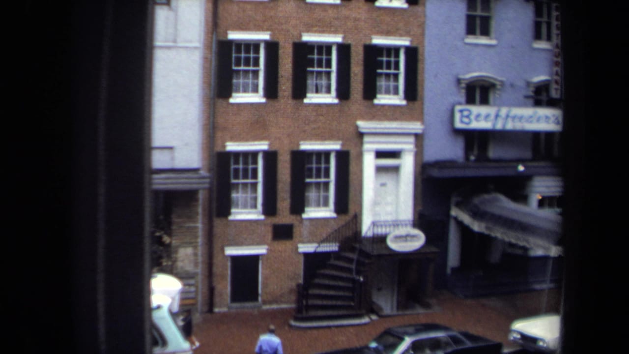 1970 Washington, DC street view with brick row buildings, black shutters, parked cars, storefront signage, and a pedestrian on the sidewalk.