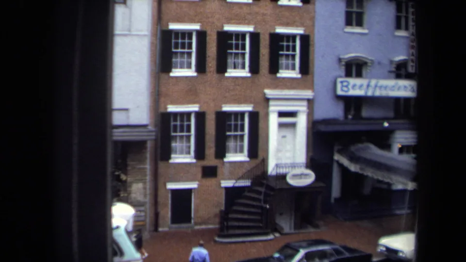 1970 Washington, DC street view with brick row buildings, black shutters, parked cars, storefront signage, and a pedestrian on the sidewalk.